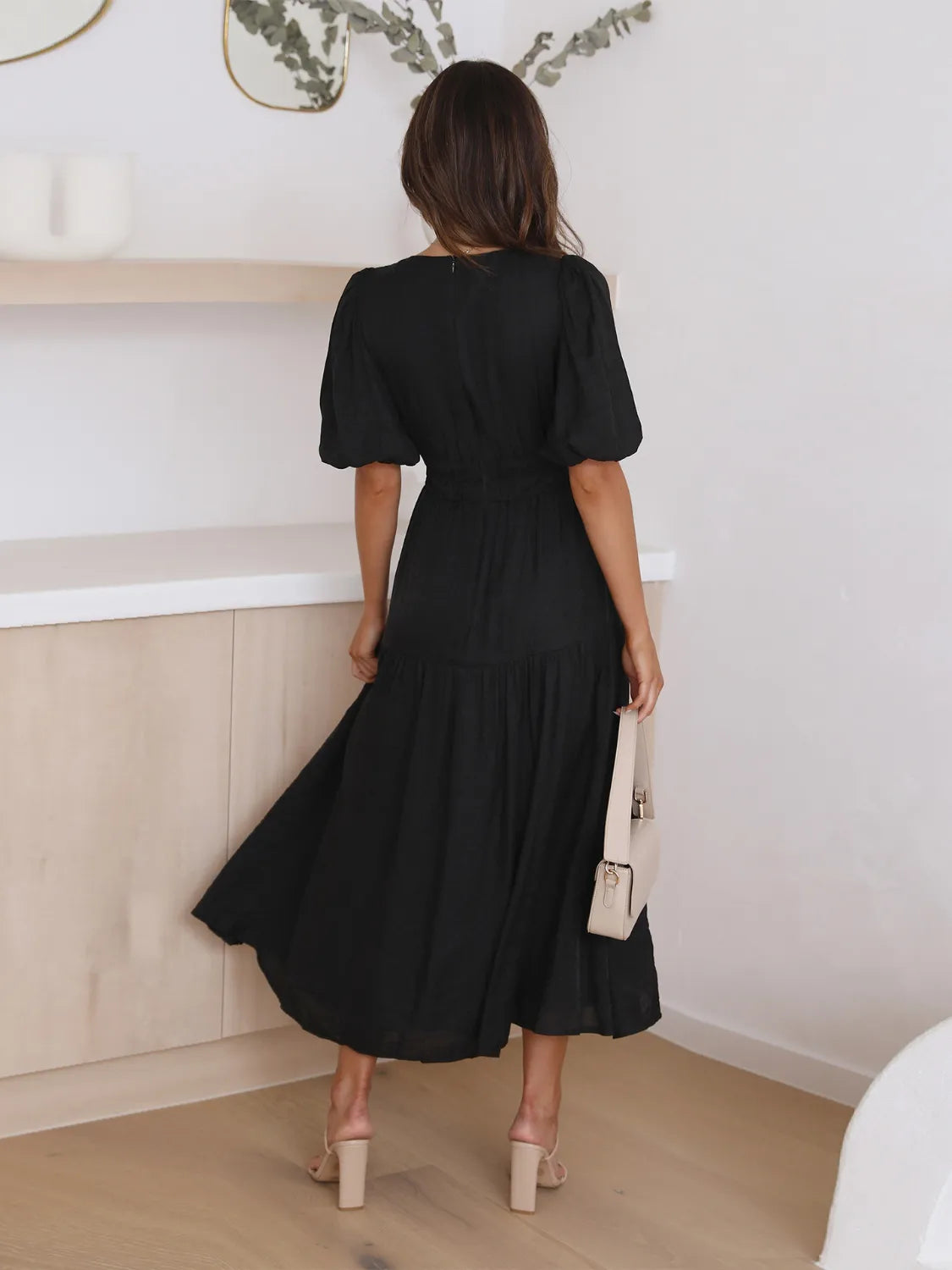 Tiered surplice half-sleeve black midi dress, back view; paired with beige heeled sandals and a beige handbag.