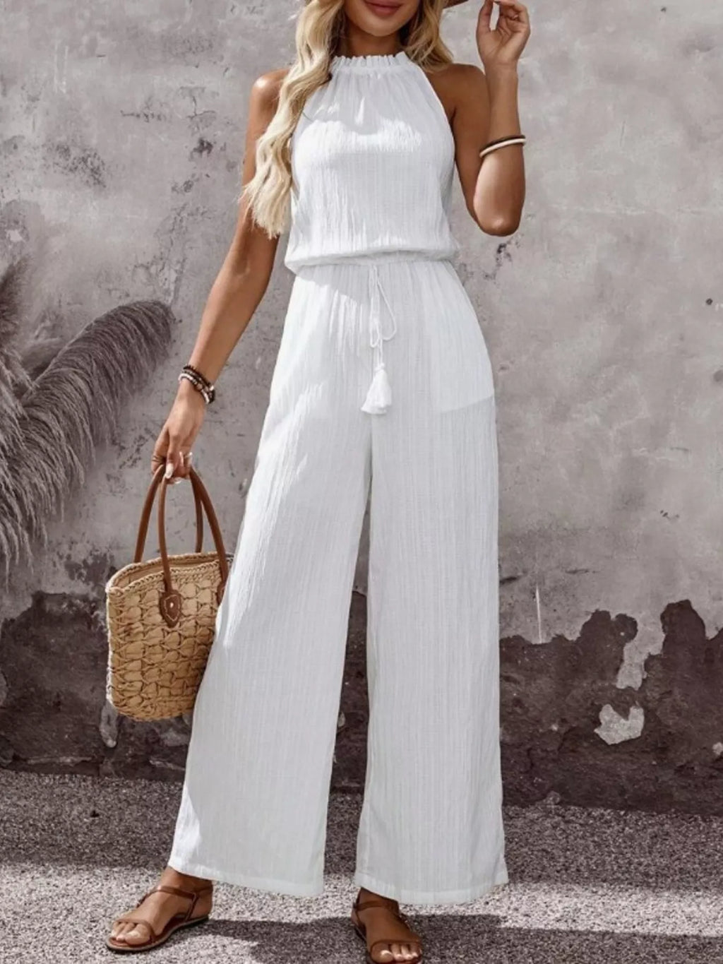 Plus-size white tassel-waist halter-neck sleeveless wide-leg jumpsuit with drawstring, paired with a straw handbag.