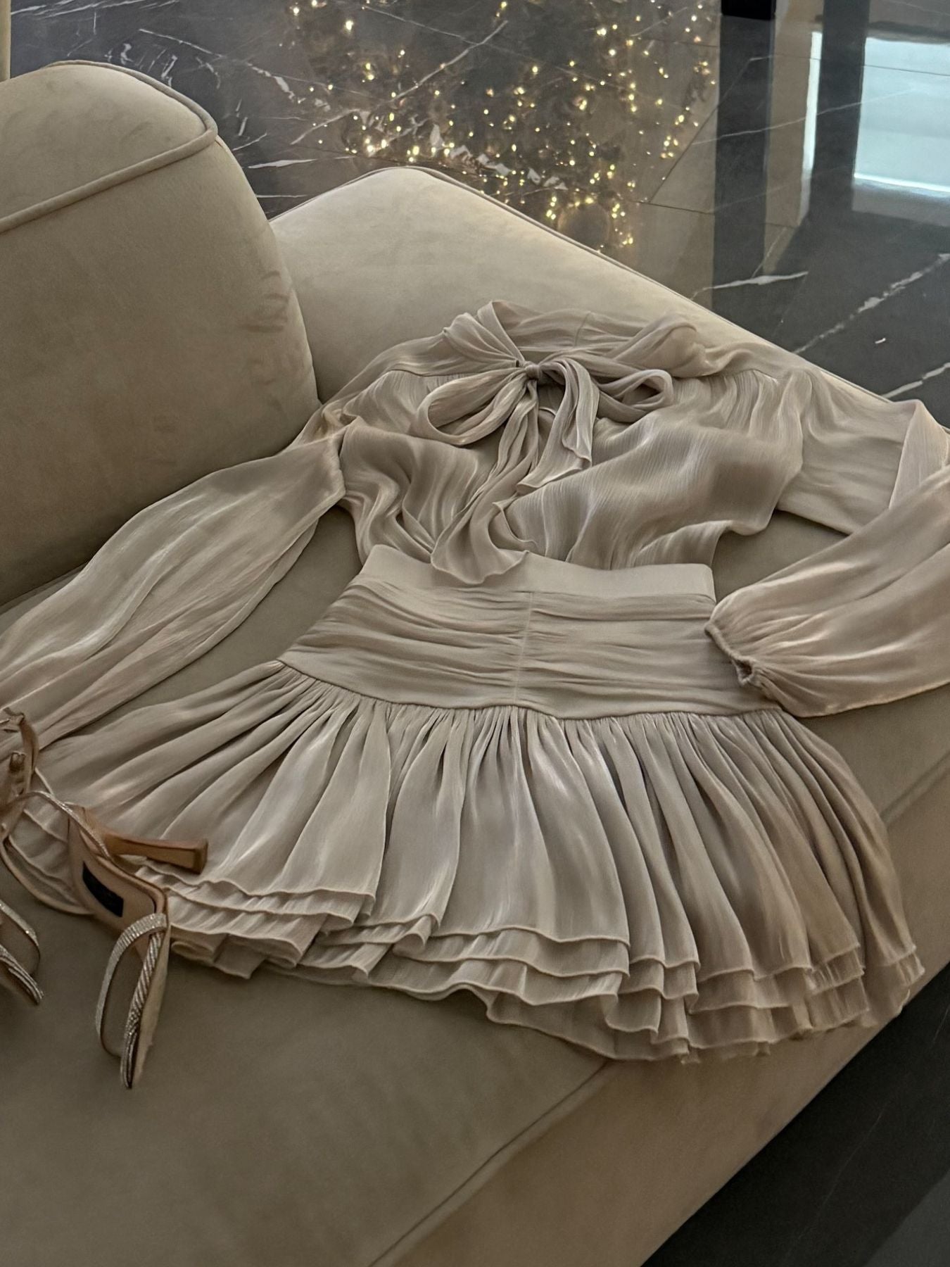 Bow-Tie Blouse and Ruched Mini Skirt Set by Trendsi in cream-beige, pleated skirt shown on a sofa