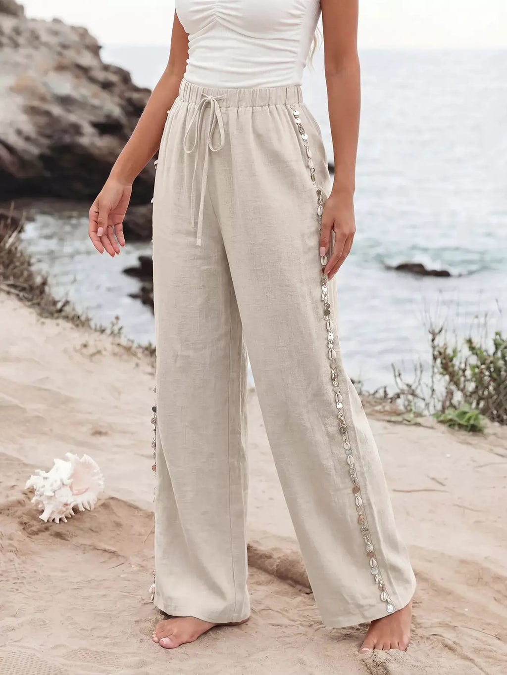 Drawstring wide-leg beige linen-look pants with a tied waist and decorative silver chain trim along the sides.